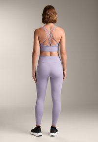 Komplet športne obleke v svetlo vijolični barvi, ki vključuje modrček z zasnovo križnih naramnic na hrbtni strani in dolge legice z gladko teksturo.