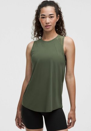 Donna con capelli ricci che indossa una canotta verde oliva e pantaloncini neri, in piedi su uno sfondo chiaro e semplice.