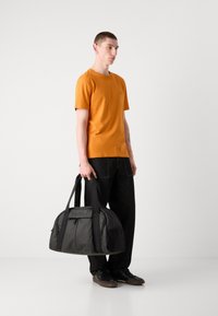 Sort polstret duffelbag med to håndtag og logodetalje, parret med en model i en orange t-shirt og mørke jeans.