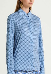 Camicia a maniche lunghe blu con motivo geometrico, dotata di colletto e bottoni, realizzata in un tessuto liscio. Gli orecchini sono color oro.