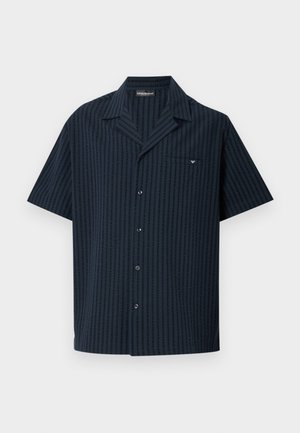 Homme grand debout pieds nus portant une chemise noire à manches courtes déboutonnée et un short de bain bleu marine avec un motif de logo blanc sur un sol gris.