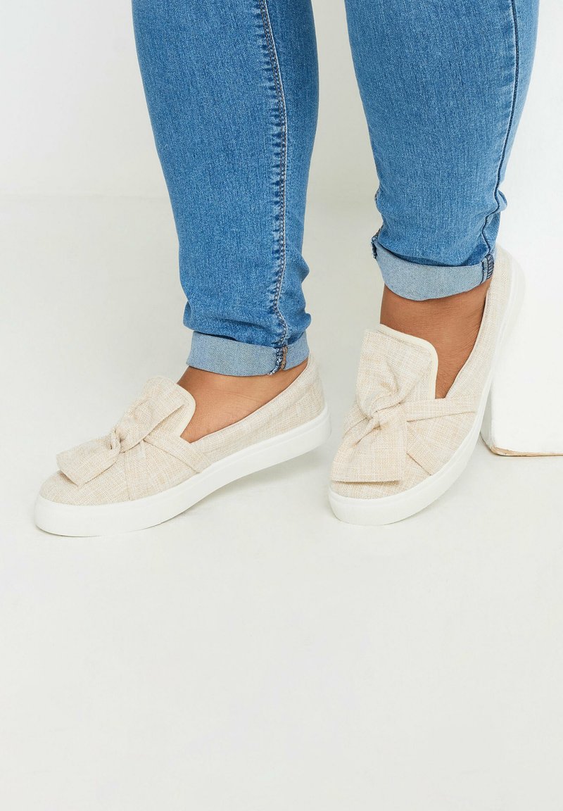 Scarpe slip-on in tessuto beige con un dettaglio a fiocco sulla parte anteriore, dotate di una suola bianca e una finitura testurizzata. Indossate con jeans blu svasati.