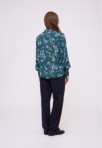 Langärmlige Bluse in tiefem Türkis mit einem floralen Muster in Blau- und Cremetönen. Kombiniert mit dunklen Hosen. Rückansicht des Outfits.
