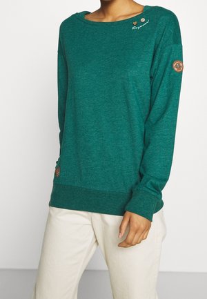 Pull bleu sarcelle à manches longues avec encolure bateau, bouton et détail en forme de cœur près de l'épaule, patchs circulaires marron sur la manche et l'ourlet, porté avec un pantalon clair.