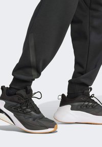 Black athletic shoes feature textured overlays, a white sole, and black laces. Worn with black tapered sweatpants and a textured ankle area.