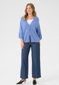 Cardigan bleu clair avec fermeture à bouton, poignets côtelés et coupe décontractée, associé à un jean en denim foncé à jambes larges et des mules noires.