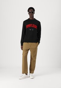 Svart strikket genser med rød "TOMMY JEANS"-logo, kombinert med brune bukser og hvite sneakers, båret av en mann med solbriller.