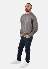 Camisa gris de manga larga con cuello redondo, jeans azul oscuro y zapatillas blancas. La camisa tiene un corte relajado y puños acanalados.