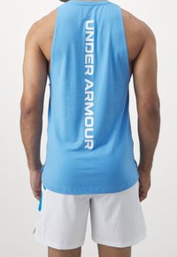 Top girocollo senza maniche blu con la scritta "UNDER ARMOUR" stampata verticalmente in bianco sulla schiena. Tessuto morbido con una vestibilità rilassata, abbinato a pantaloni corti bianchi.