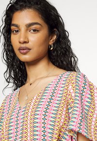 Top colorido e estampado com um decote em V, apresentando designs em zig-zag nas cores rosa, amarelo e azul. Brincos de argola dourados e colares em camadas são visíveis.