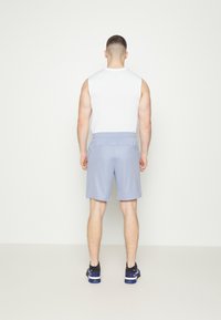 Moda masculina: top blanco sin mangas, pantalones cortos azul claro con cinturilla elástica, diseño minimalista. Zapatillas negras con detalles azules y suelas texturizadas.