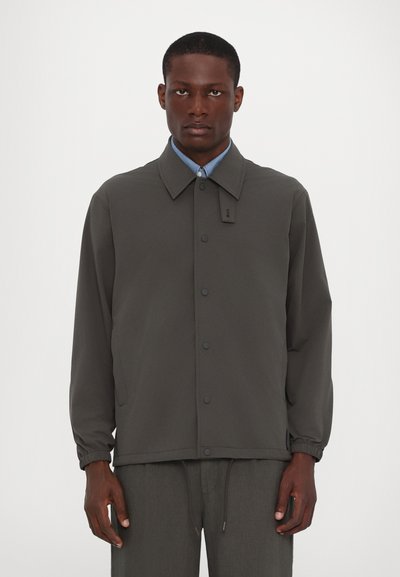 Dunkelgraue Jacke mit kragenförmigem Ausschnitt, vorderer Druckknopfverschlüsse, elastischen Bündchen und strukturiertem Stoff. Getragen über einem hellblauen Hemd.