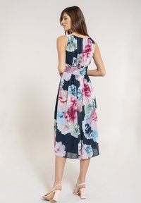 Frau in einem ärmellosen, marineblauen Kleid mit großen pinken, blauen und grünen Blumenmustern, die mit Händen in den Hüften vor einem weißen Hintergrund steht.