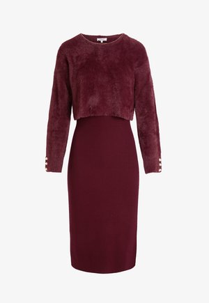 Maglione corto in pelliccia sintetica di colore bordeaux, con maniche lunghe e dettagli con bottoni. Gonna aderente a coste bordeaux al di sotto. Texture liscia su tutta la superficie.