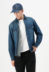 Jeune homme portant une veste en denim bleu sur une chemise blanche, une casquette à motif bleu et un pantalon noir, posant devant un fond blanc.