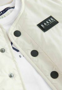 Gepolsterte cremefarbene Jacke mit einem schwarzen Logo-Patch, ausgestattet mit Druckknöpfen und einer glatten Textur, über einem weißen Hemd mit marineblauen Details getragen.