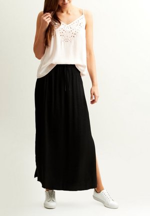 Vrouw die een witte mouwloze top met borduurwerk draagt, lange zwarte rok met een zijsplit en witte sneakers, staand tegen een effen achtergrond.