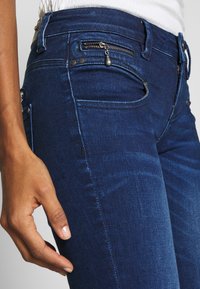 Vänsterhanden vilar nära låret på en person som bär mörkblå skinny jeans med synliga framfickor och en liten dragkedjeficka.