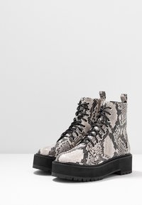Botas de plataforma gruesas con cordones y suelas negras, que presentan un patrón de serpiente en gris y negro, sobre un fondo blanco liso.