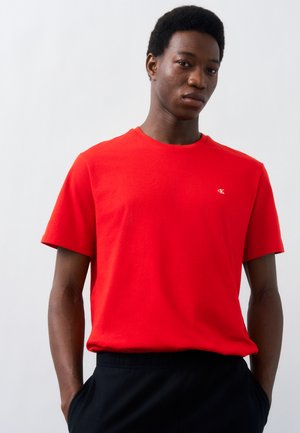 Mladý muž s krátkými kudrnatými vlasy, oblečený v červeném tričku Calvin Klein a černých kalhotách, stojící na světlém neutrálním pozadí.