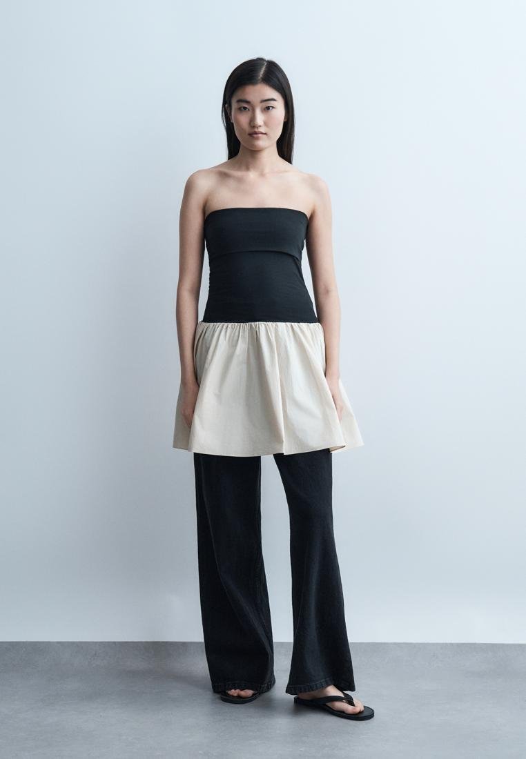 Femme debout contre un fond uni portant un haut bustier noir et beige, un pantalon large noir et des tongs noires.