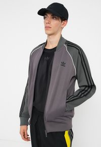 Siva i tamnosiva jakna na zip s bijelim detaljima i tri crne pruge na rukavima, nošena preko crne majice. Crna kapa s logom.