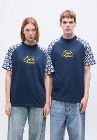 Μπλε ναυτικά μπλουζάκια με κοντά μανίκια με σχέδιο, το κεντημένο λογότυπο «Carlo Olucci» σε κίτρινο χρώμα. Χαλαρή εφαρμογή, συνδυασμένα με ανοιχτό μπλε τζιν.