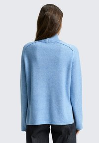 Hellblauer Strickpullover mit einem Stehkragen, geripptem Rand und überschnittenen Schultern. Weiche Textur und lockere Passform, kombiniert mit dunklen Hosen.