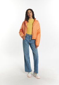 Jachetă puf orange, pulover galben cu dungi, blugi deschis-colorați cu picioare largi și adidași multicolori. Păr creț, mâinile în buzunare.