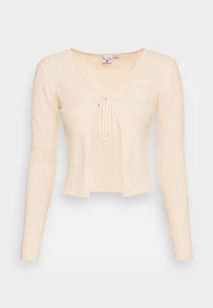 Beige ribbattu cropped neule, jossa on pitkät hihat, syvä V-pääntie, solmittava etusulku ja pieni brodeerattu logo vasemmalla puolella.