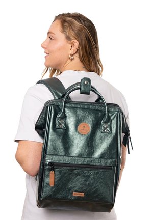 Groene metallic cosmetica rugzak met dubbele schouderbanden, handvat aan de bovenkant, ritsvak aan de voorkant en een ronde logopatch op de voorkant.