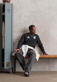 Crna dukserica s bijelim logotipom, sivi široki hlače i tenisice. Ormar u neutralnim tonovima i drvena klupa u minimalističkom okruženju.