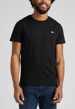 Hymyilevä mies, joka pukeutunut yksinkertaiseen mustaan Lee-t-paitaan ja sinisiin farkkuihin, seisoo vaaleanharmaan taustan edessä.