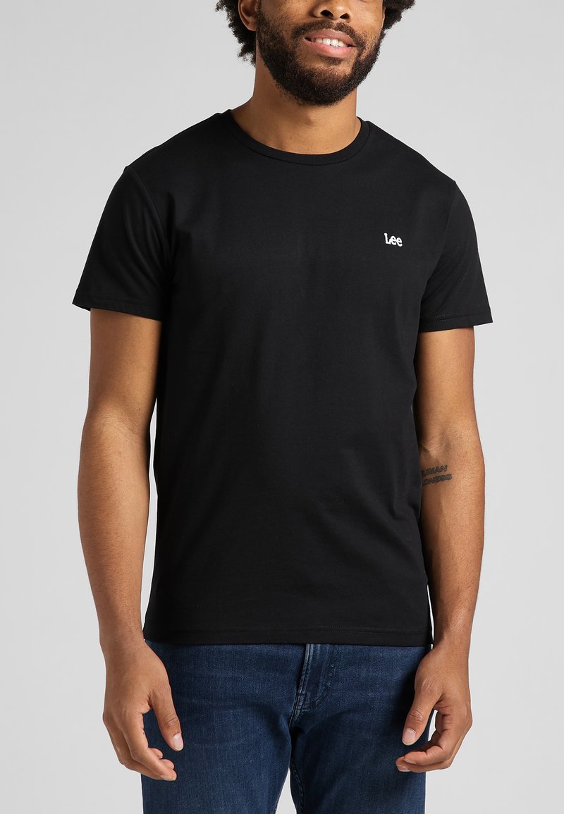 Χαμογελαστός άνδρας που φοράει μια απλή μαύρη μπλούζα Lee και μπλε τζιν, στέκεται μπροστά σε ένα ανοιχτό γκρι φόντο.