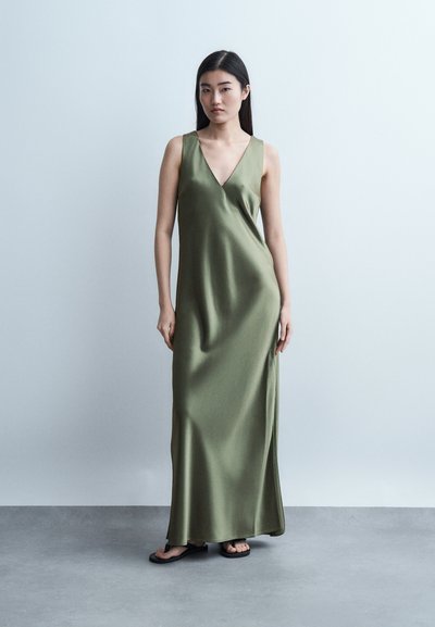 Γυναίκα που στέκεται σε μακριά αμάνικη σατέν φόρεμα σε χρώμα ελιάς με V-λαιμόκοψη, φοράει μαύρα σανδάλια, σε απλό φωτεινό γκρι φόντο.