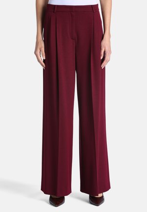 Femme portant un pantalon large bordeaux taille haute associé à des chaussures pointues foncées, debout devant un fond clair uni.