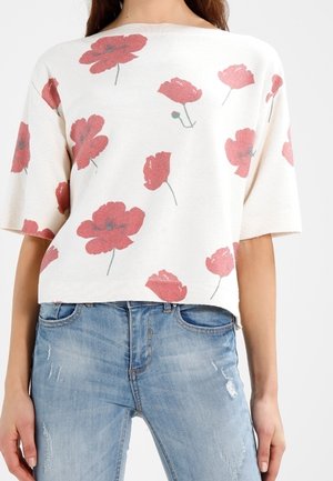 Femme portant un pull blanc à manches courtes avec des imprimés floraux rouges et un jean bleu clair usé.