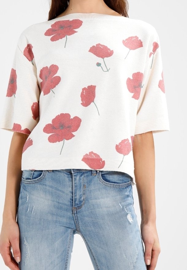 Femme portant un pull blanc à manches courtes avec des imprimés floraux rouges et un jean bleu clair usé.