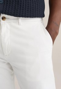 Hvide chinos med en flad front og sidelommer, der har en brun knap og en glat tekstur, kombineret med en struktureret marineblå sweater.