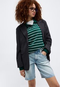 Γυναίκα με σγουρά μαλλιά, φορώντας μαύρο σακάκι, πράσινη ριγέ πόλο μπλούζα, ανοιχτόχρωμα τζιν σορτς και γυαλιά, ποζάρει με το χέρι στην τσέπη.