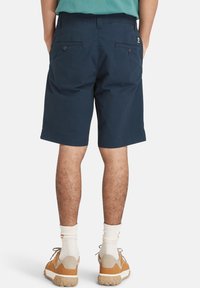 Marineblauwe shorts met een platte voorkant, achterzakken beveiligd met knopen, gemaakt van lichtgewicht stof. gedragen met lichtgekleurde sneakers en sokken.