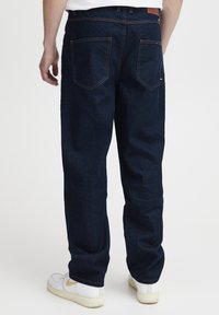 Jeans in denim blu scuro con una vestibilità dritta, dotati di due tasche posteriori e cuciture a contrasto, indossati con sneaker bianche.