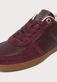 Μαρόν αθλητικό παπούτσι Tommy Hilfiger με υφή σουέτ και δέρματος, καφέ λαστιχένια σόλα και χρυσαφί branded οπές και ετικέτα στη γλώσσα.