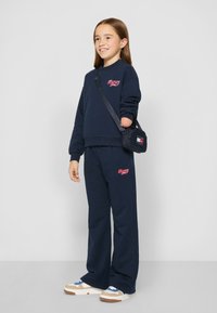 Sudadera marina con logo, pantalones de chándal marinos a juego, zapatillas deportivas casuales con acentos en beige y azul, y un pequeño bolso cruzado.