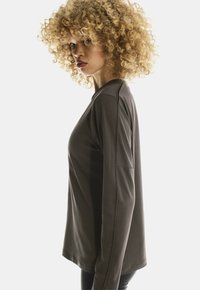 Vrouw met krullend blond haar in een langwerpig grijs shirt, zichtbaar in zijaanzicht tegen een witte achtergrond.