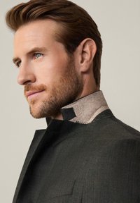 Giacca scura con texture e colletto alto; il colletto presenta un morbido accento beige e nero. Capelli lisci, primo piano di profilo.