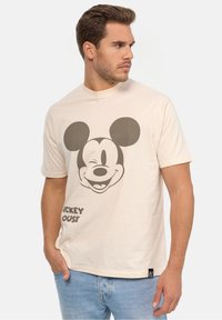 Beige puuvillatee, jossa on suuri mustavalkoinen Mickey Mouse -grafiikka ja "MICKEY HOUSE" -teksti tummalla paksulla fontilla. Klassinen malli.