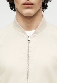 Κοντινό πλάνο ενός ατόμου που φοράει μπεζ μπουφάν τύπου bomber με ριμπ ύφασμα στο γιακά και ασημένιο φερμουάρ μπροστά, με εμφανή τα χείλη και το πηγούνι, φόντο καθαρό λευκό.