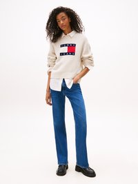 Beige knitted sweater with blue, white, and red logo patch, worn over a white collared shirt. High-waisted blue jeans and black chunky shoes.