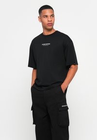 Zwart oversized T-shirt gemaakt van katoen, met een ronde hals en de tekst "STUDIO EDITION" op de borst. Gepaarde met zwarte cargo broeken.